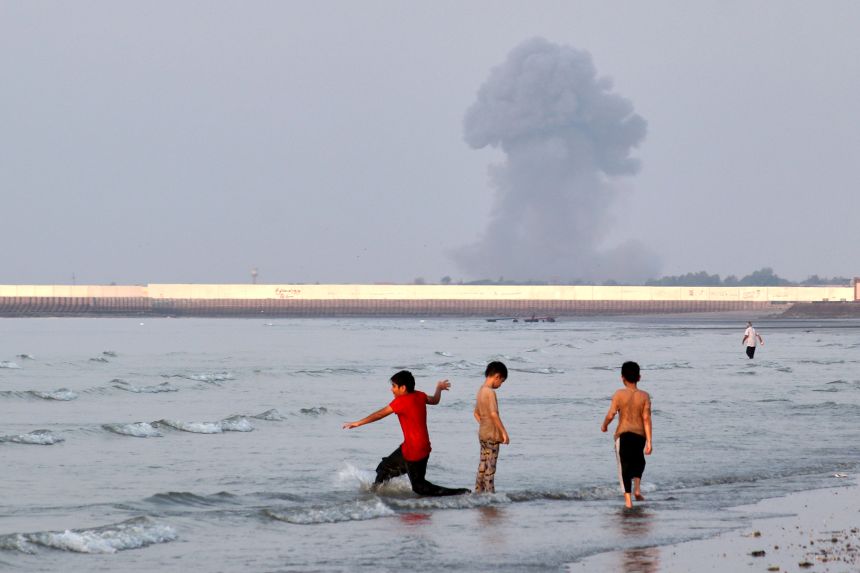 Three boys play in the shallow waters of the Strait of Hormuz, as a plume of smoke rises from an explosion in the background, off Bandar Abbas, Iran, Monday, July 13, 2026. (Razieh Poudat/ISNA via AP)